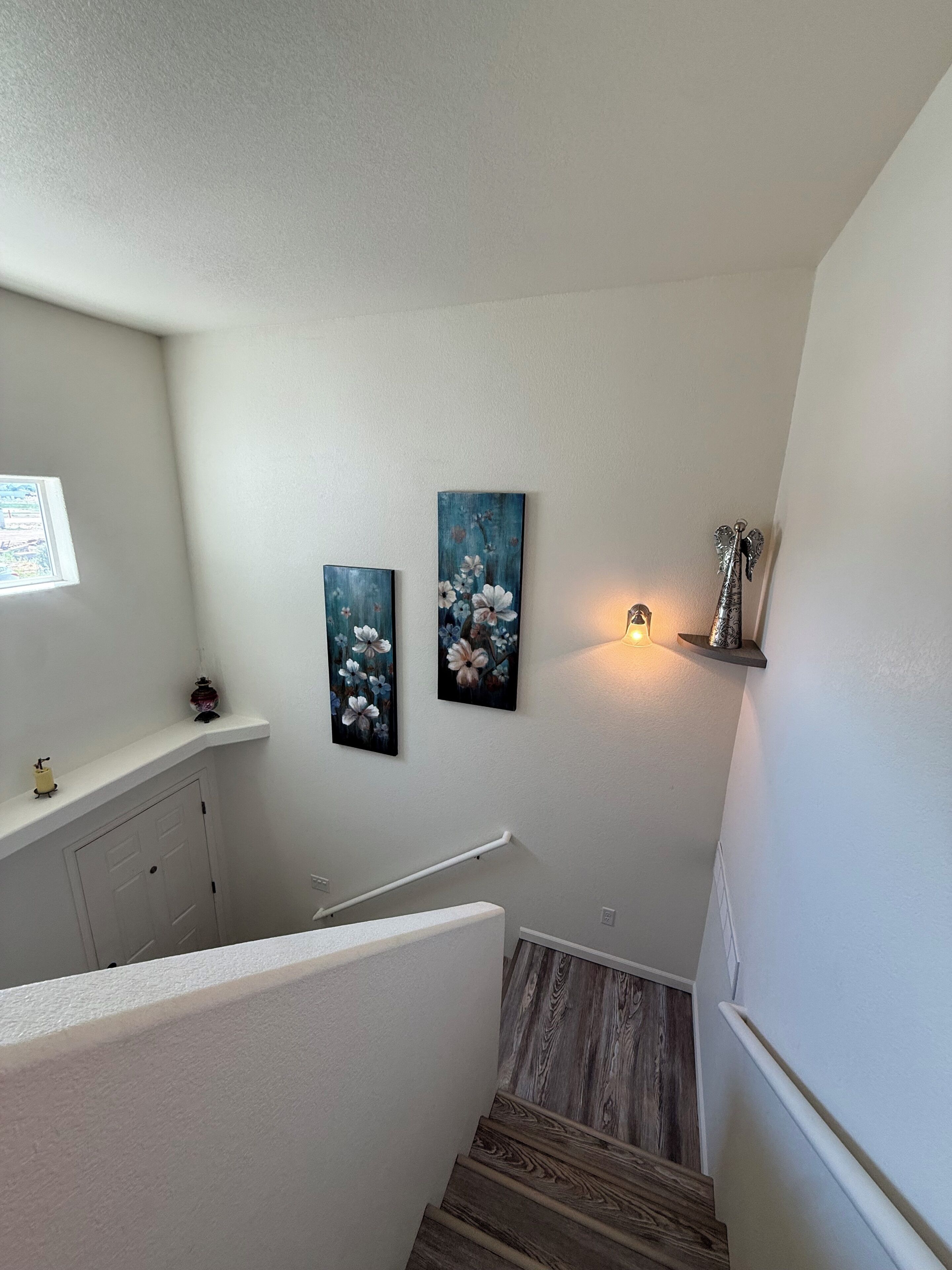 Una escalera con barandilla, un estante adosado a la pared con adornos, y dos cuadros enmarcados con motivos florales.