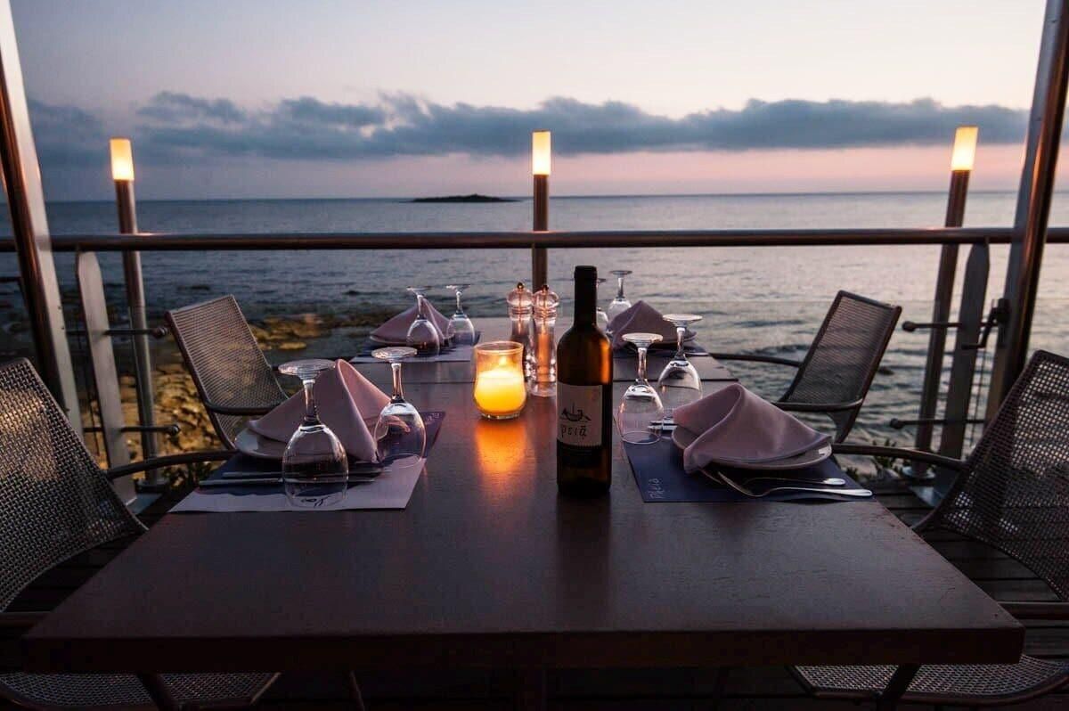 A table set for a meal with a candle, wine bottle, and glasses, overlooking a body of water.