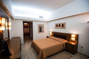 Habitación de hotel con cama, mesitas de noche, escritorio y espejo. Se ve una puerta que da a otra habitación.