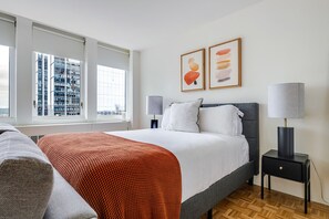 A hotel room with a bed, two bedside lamps, a window with a view of buildings, and two framed artworks on the wall. - Blueground | Prudential / St. Botolph, view, nr Boston Public Garden (Boston)