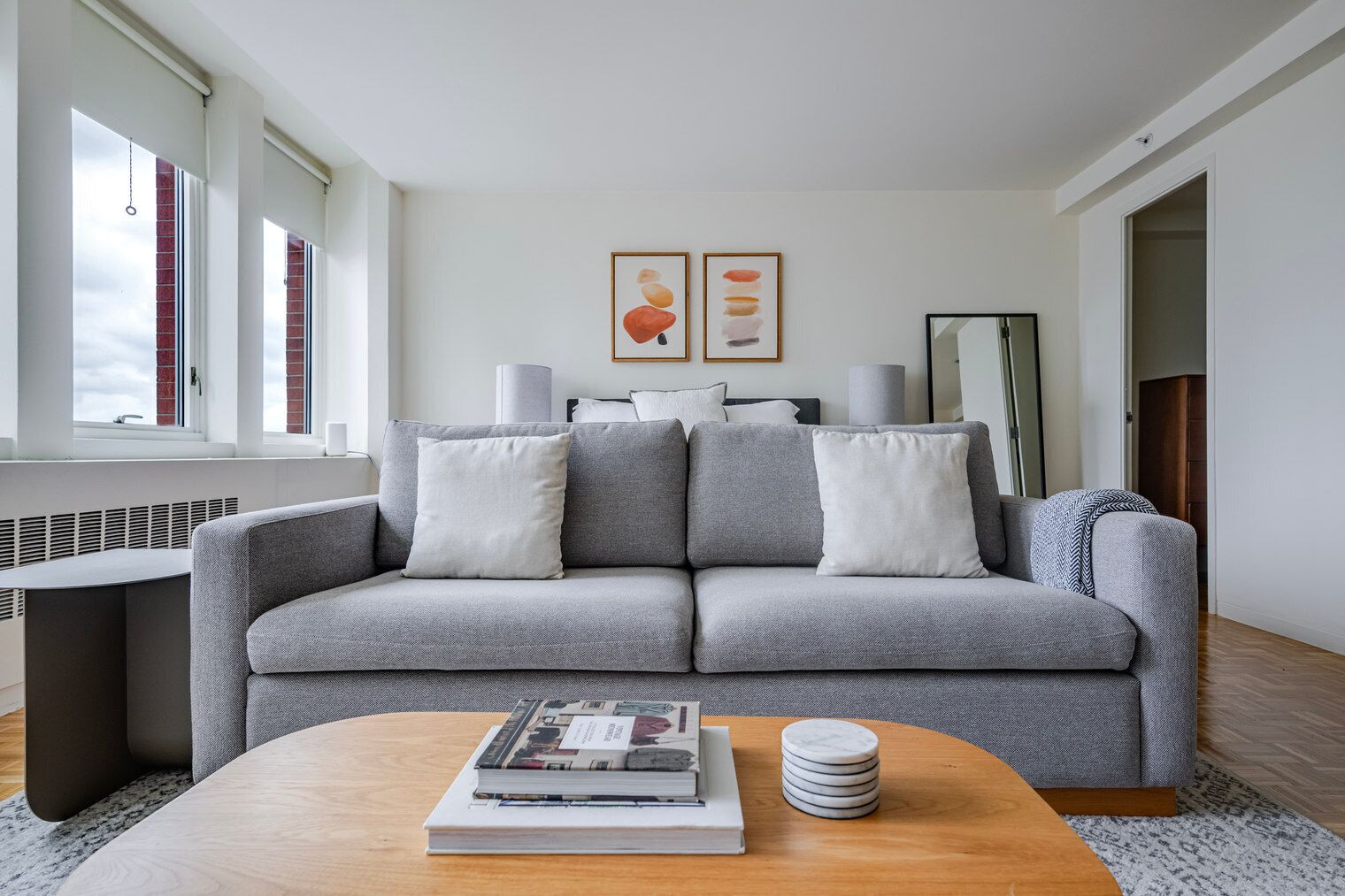 Ein modernes Wohnzimmer mit einem grauen Sofa, einem Couchtisch mit Büchern und Untersetzern sowie einem Fenster mit Jalousien.