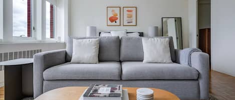 Una sala de estar moderna con un sofá gris, una mesa de centro con libros y posavasos, y un ventanal con persianas.