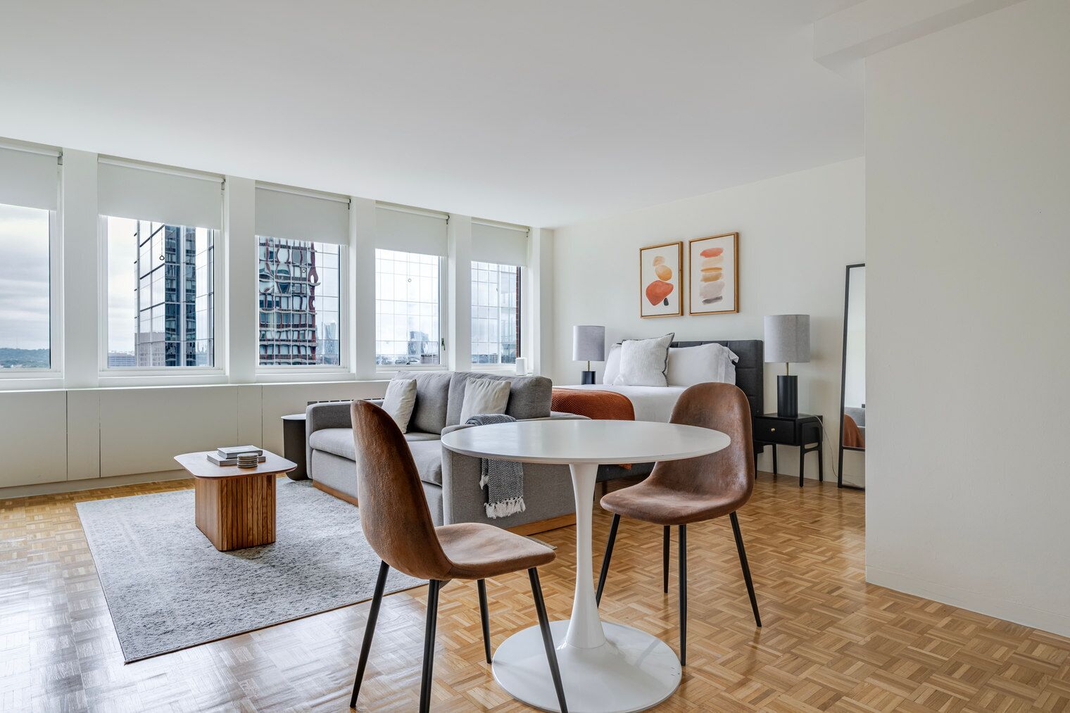 A modern hotel room with a large window, a sofa, a dining table with chairs, and a bed.