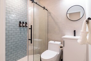 A bathroom with a glass shower enclosure, a white toilet, a sink with a mirror, and toiletries on the wall. - Thena Hotel - Beautiful 1 Bedroom (Philadelphia)