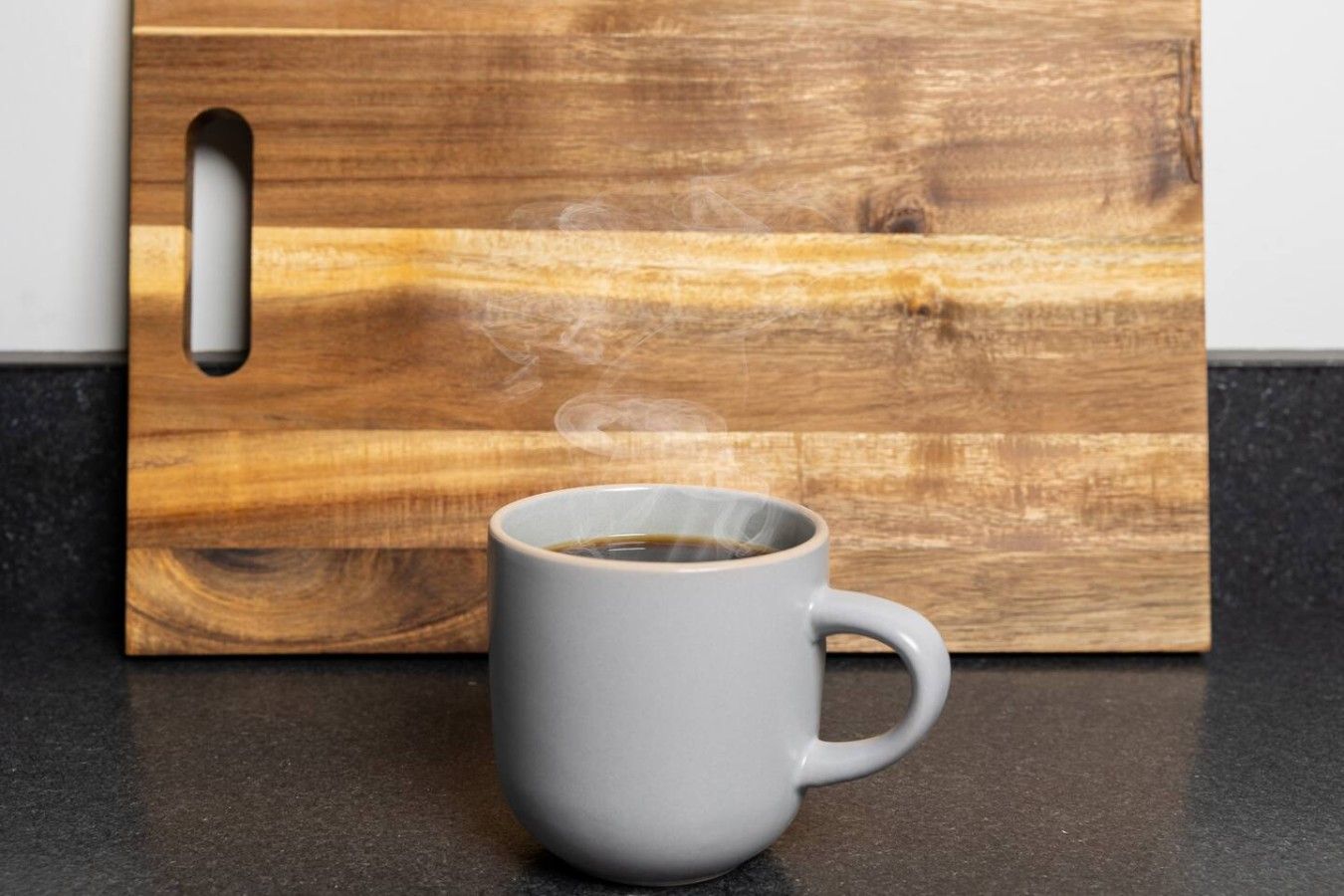 Une tasse blanche posée sur un plan de travail foncé, de laquelle s’échappe de la vapeur, et placée devant une planche à découper en bois dotée d’une poignée.