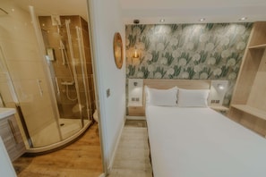 Habitación de hotel con ducha envuelta en vidrio, cama con ropa blanca y pared con estampado de hojas.