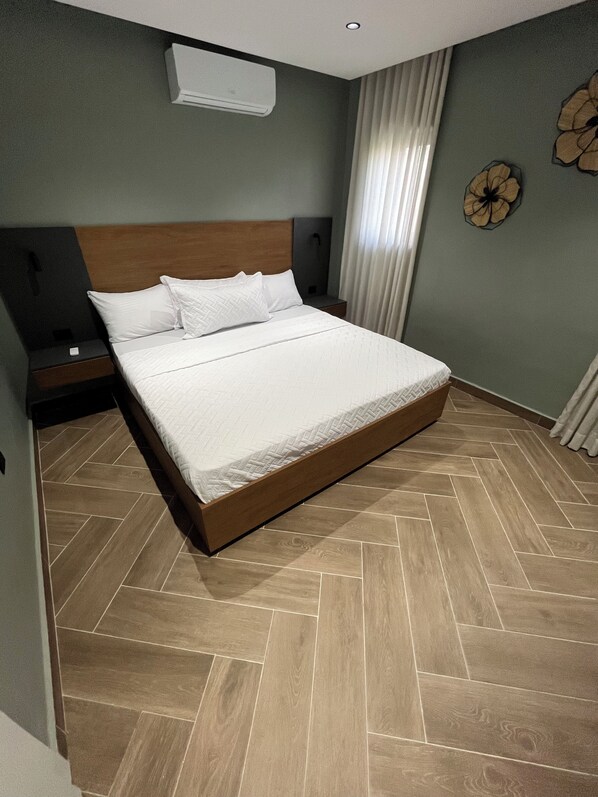 A hotel room with a large bed, a nightstand, a wall-mounted air conditioner, and a decorative wall piece. - Vrbo Property (Higuey)