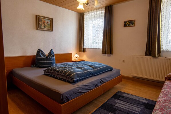 Een enkel bed met houten hoofdeinde, een nachtkastje met een lamp en een raam met gordijnen.