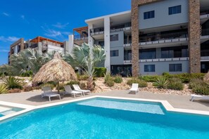 A swimming pool with lounge chairs and a thatched umbrella, surrounded by a landscaped garden with palm trees and cacti. - Apartment - Perla di Cape Marie 1412 (Jan Kok)