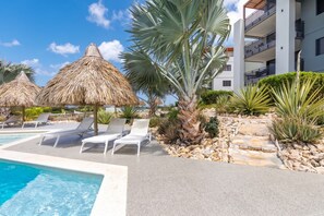 A pool area with lounge chairs and thatched umbrellas, surrounded by tropical plants and a building with balconies. - Ground-Floor 2BR Condo · Pool steps away · Curaçao (Jan Kok)