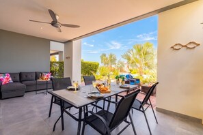 An open-plan living area with a dining table set for four, a ceiling fan, and a view of a garden with palm trees. - Apartment - Perla di Cape Marie 1412 (Jan Kok)