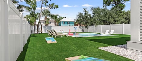 A backyard with artificial grass, a pool, lounge chairs, and a wooden deck.