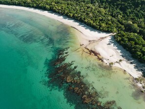 Luchtbeeld van een strand met helder turquoise water, een wit zandstrand en een dichtgroen bos.