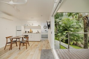 Een moderne keuken met witte kasten, een eetgedeelte met houten stoelen en uitzicht op een weelderige tropische tuin door een glazen deur.