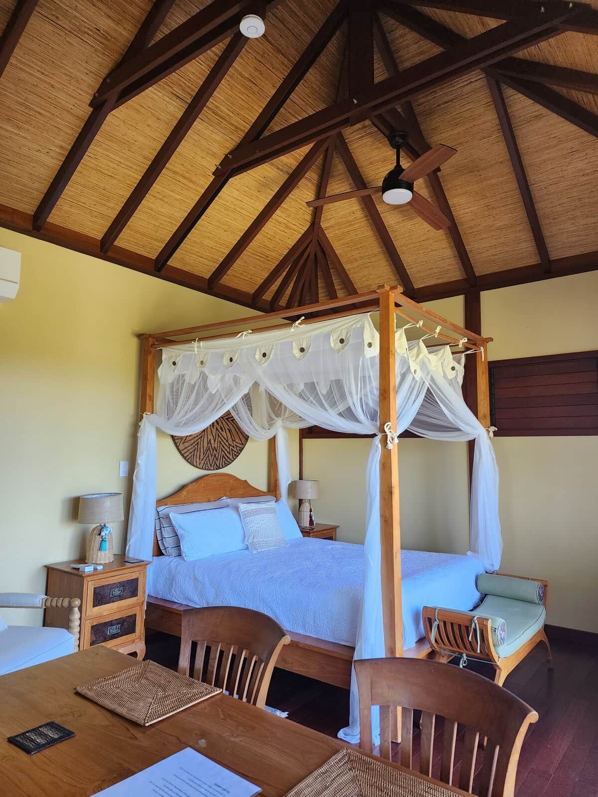 A four-poster bed with a canopy, a wooden table with chairs, and a ceiling fan.