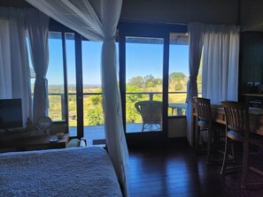 Un dormitorio con una cama grande, un escritorio y una silla, con vista a un balcón y follaje exterior.