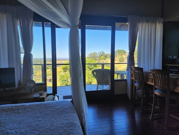 A bedroom with a large bed, a desk, and a chair, with a view of a balcony and greenery outside.