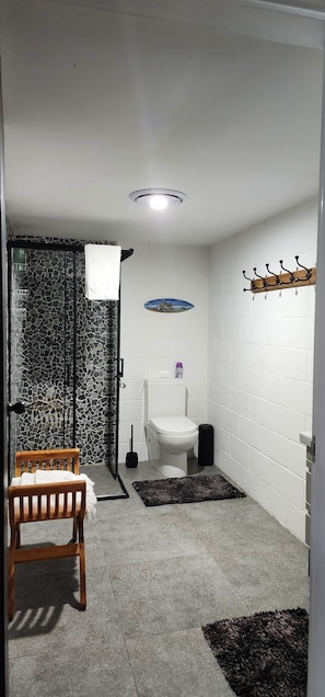 Une salle de bain comprenant des toilettes, un espace douche, une chaise en bois, un tapis de sol noir et un luminaire au plafond.