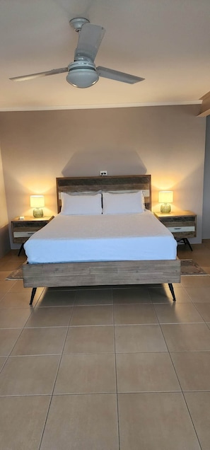 Une chambre à coucher avec un lit, deux tables de chevet avec des lampes, un ventilateur de plafond et un sol carrelé.