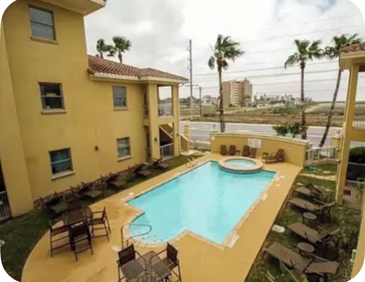 A swimming pool with lounge chairs and a small hot tub, surrounded by a building with multiple windows and a view of palm trees and buildings in the distance.