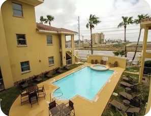 A swimming pool with lounge chairs and a small hot tub, surrounded by a building with multiple windows and a view of palm trees and buildings in the distance. - 2-bedroom/ 3 bed condo at SouthPadre Island (South Padre Island)