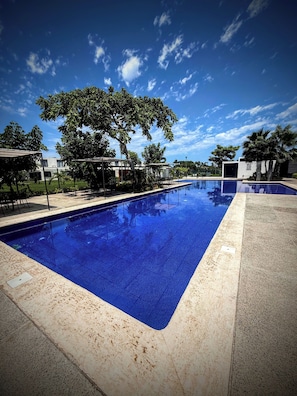 A rectangular swimming pool with a clear blue water surface, surrounded by a concrete deck and shaded by trees. - Pool-Vacation-Jobs-Grills - PUERTO CARMA (Las Juntas)