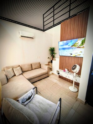 A living room with a beige sofa, a small table, a standing fan, and a TV mounted on a wooden panel. - Pool-Vacation-Jobs-Grills - PUERTO CARMA (Las Juntas)