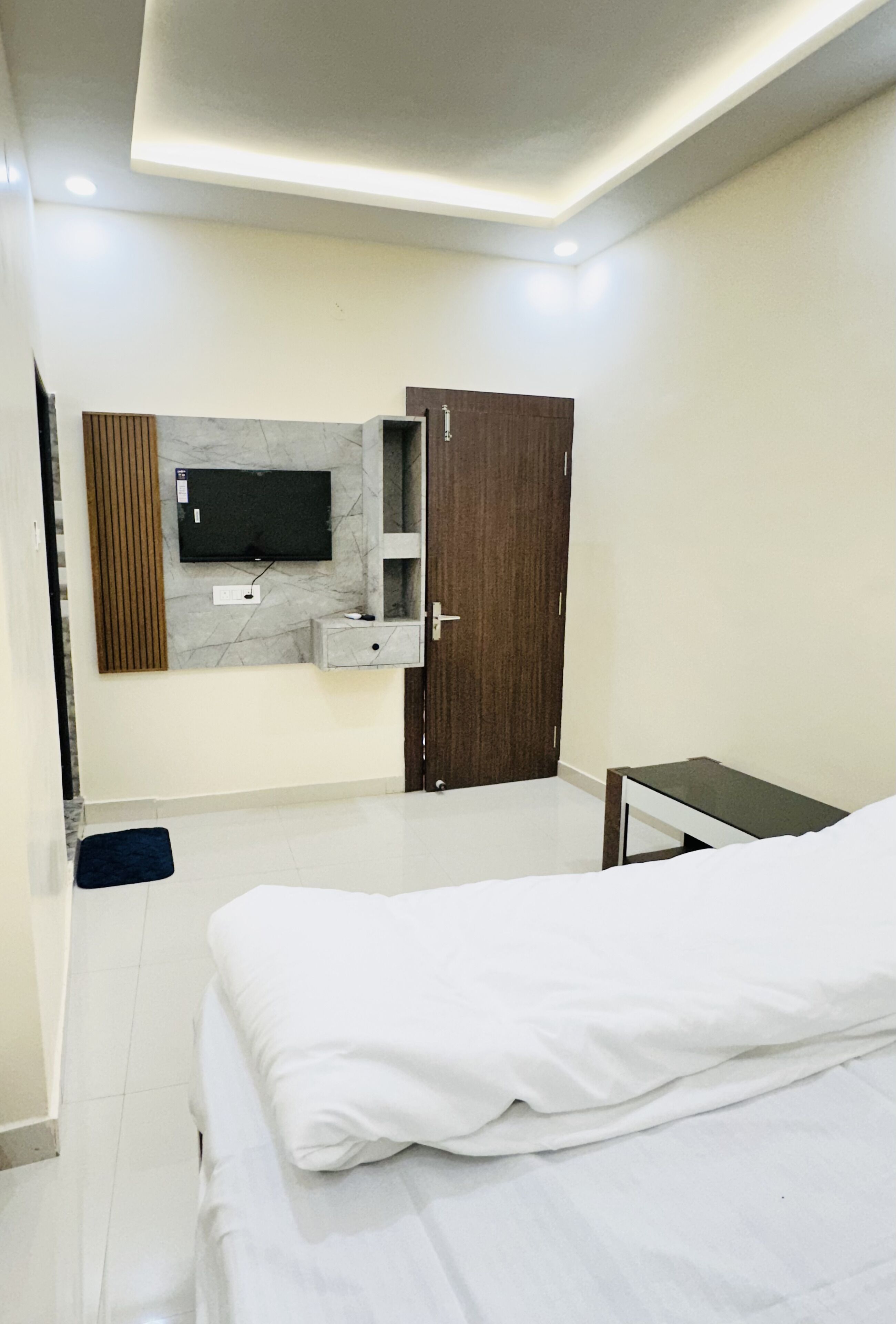 A modern bedroom with a bed, a TV mounted on the wall, a wooden wardrobe, and a small bedside table.