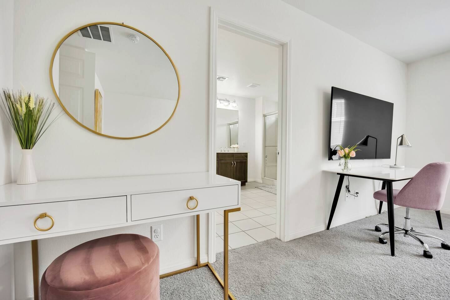 A modern bedroom with a white dresser, a round mirror, a desk with a chair, and a television.
