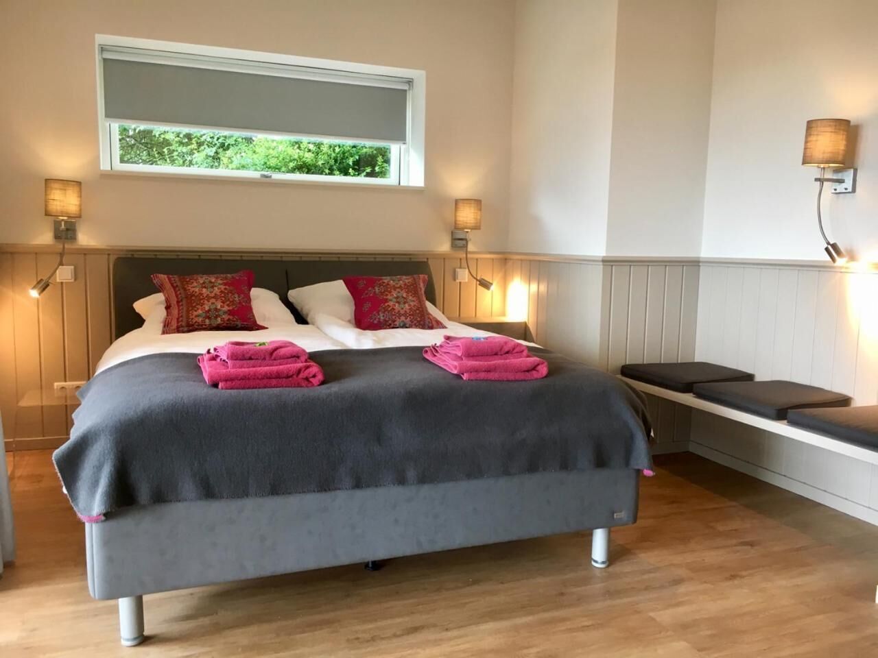 Cama bem arrumada com almofadas e toalhas dobradas, mesa de cabeceira e um banco com luminária.
