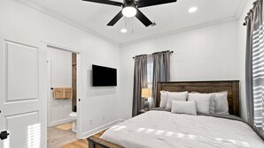 Ein Schlafzimmer mit einem Bett, einem Deckenventilator, einem an der Wand montierten Fernseher und einem Fenster mit Vorhängen.