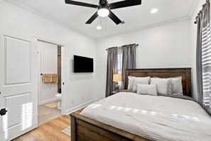 Ein Schlafzimmer mit einem Bett, einem Deckenventilator, einem an der Wand montierten Fernseher und einem Fenster mit Vorhängen.