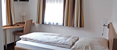 A hotel room with a single bed, a desk, a chair, a television mounted on the wall, and a window with curtains.