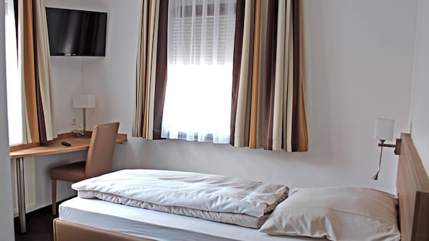 Une chambre d’hôtel avec un lit simple, un bureau, une chaise, un téléviseur fixé au mur et une fenêtre avec des rideaux.