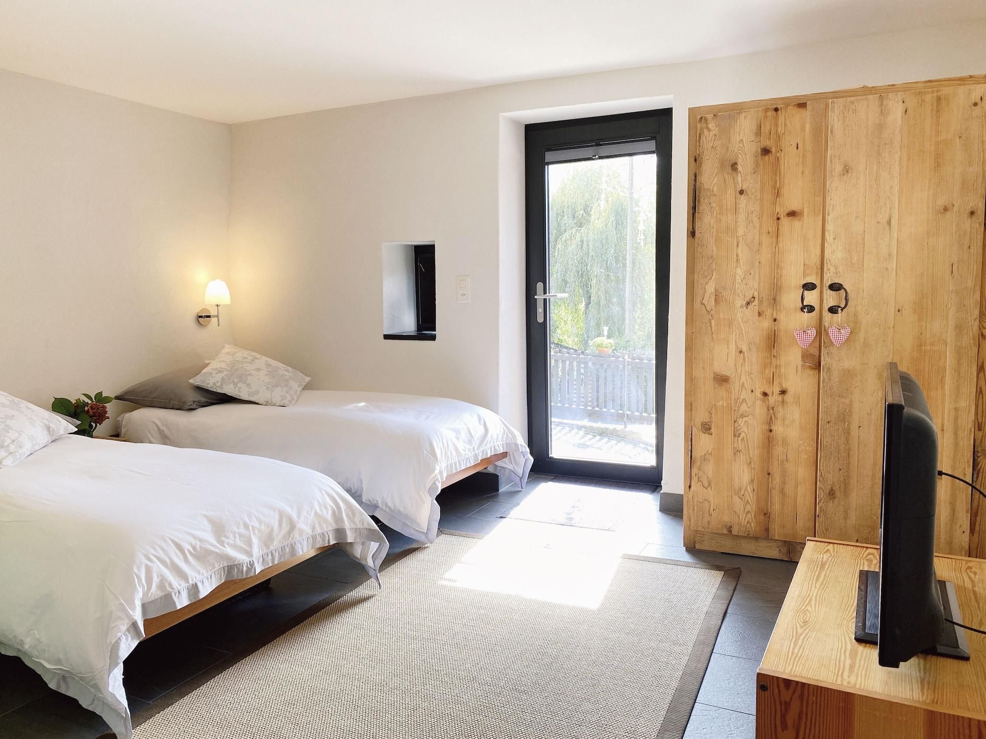 A bedroom with two beds, a wooden wardrobe, a television, and a window with a view of greenery.