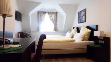 Ein Hotelzimmer mit einem Bett, einem Schreibtisch, einem Stuhl, einer Lampe, einem Bild an der Wand und einem Fenster mit Vorhängen.