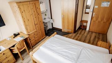 Een kamer met een bed, een houten garderobe, bureau met stoel en een klein badgedeelte.