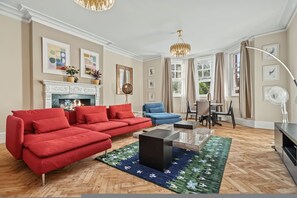 A living room with a fireplace, red sofa, blue armchair, and a dining area with a table and chairs. - Holland Park 5BR 3BA Stay With Private Sauna (London)