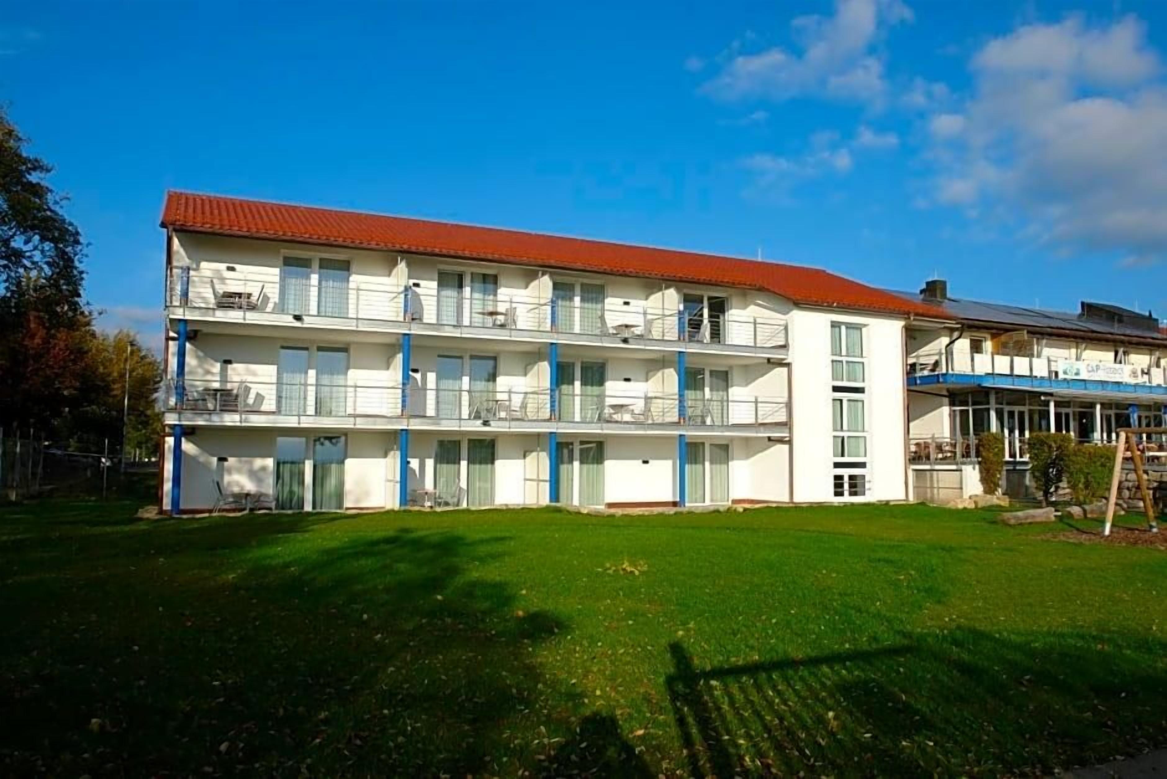 A multi-story building with balconies, a red roof, and a grassy lawn in front.