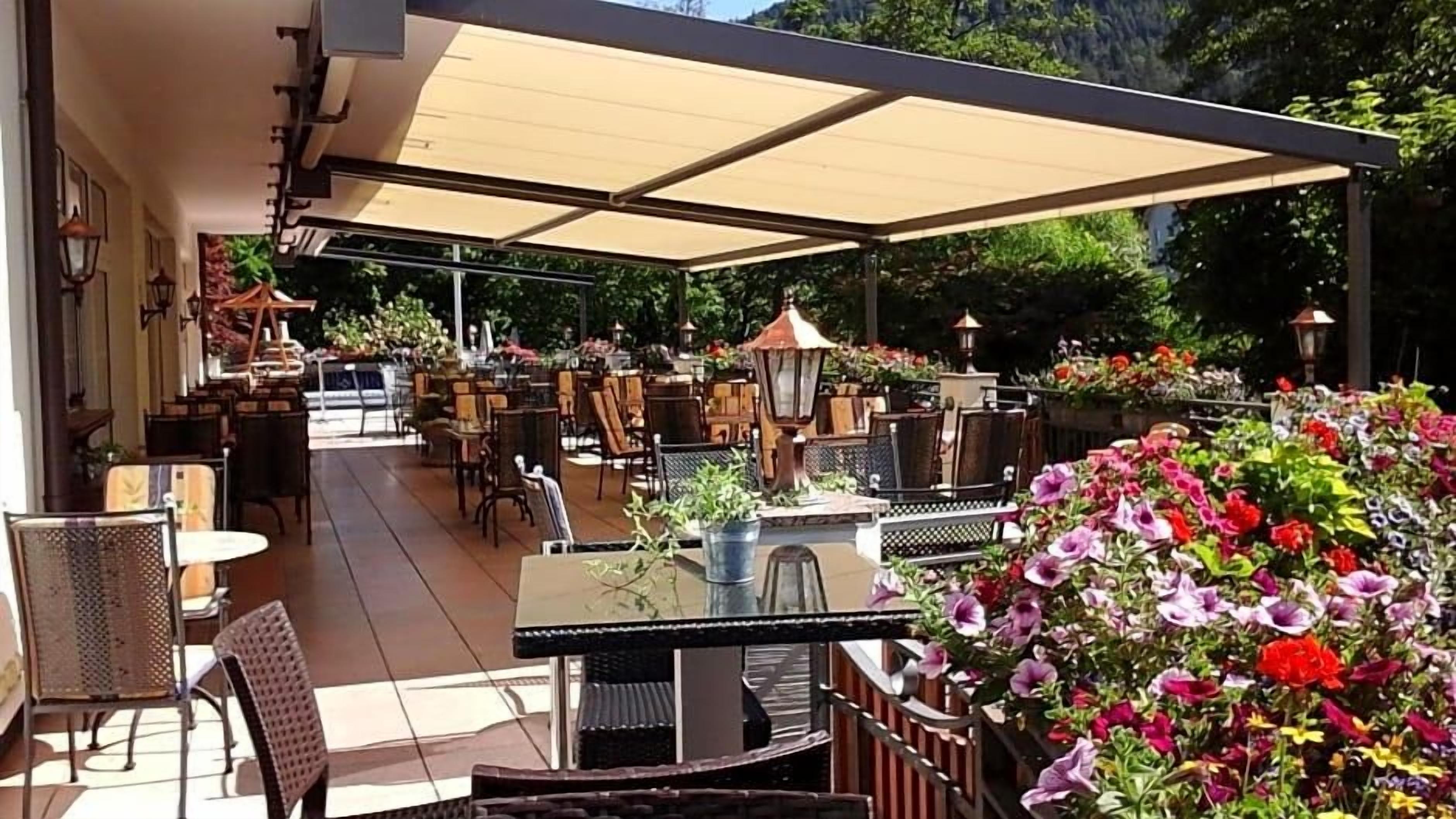 An outdoor patio area with tables and chairs, surrounded by a covered seating area and potted plants.