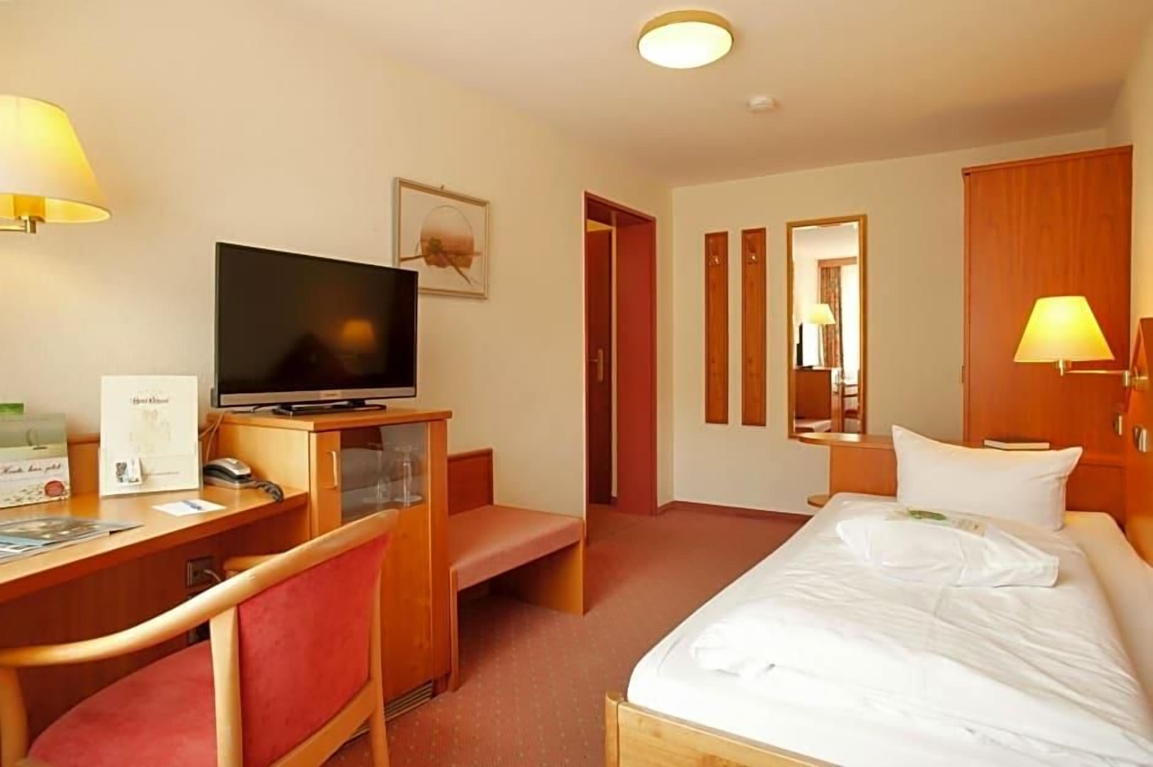 A hotel room with a bed, a desk with a television, a chair, and a bedside table.
