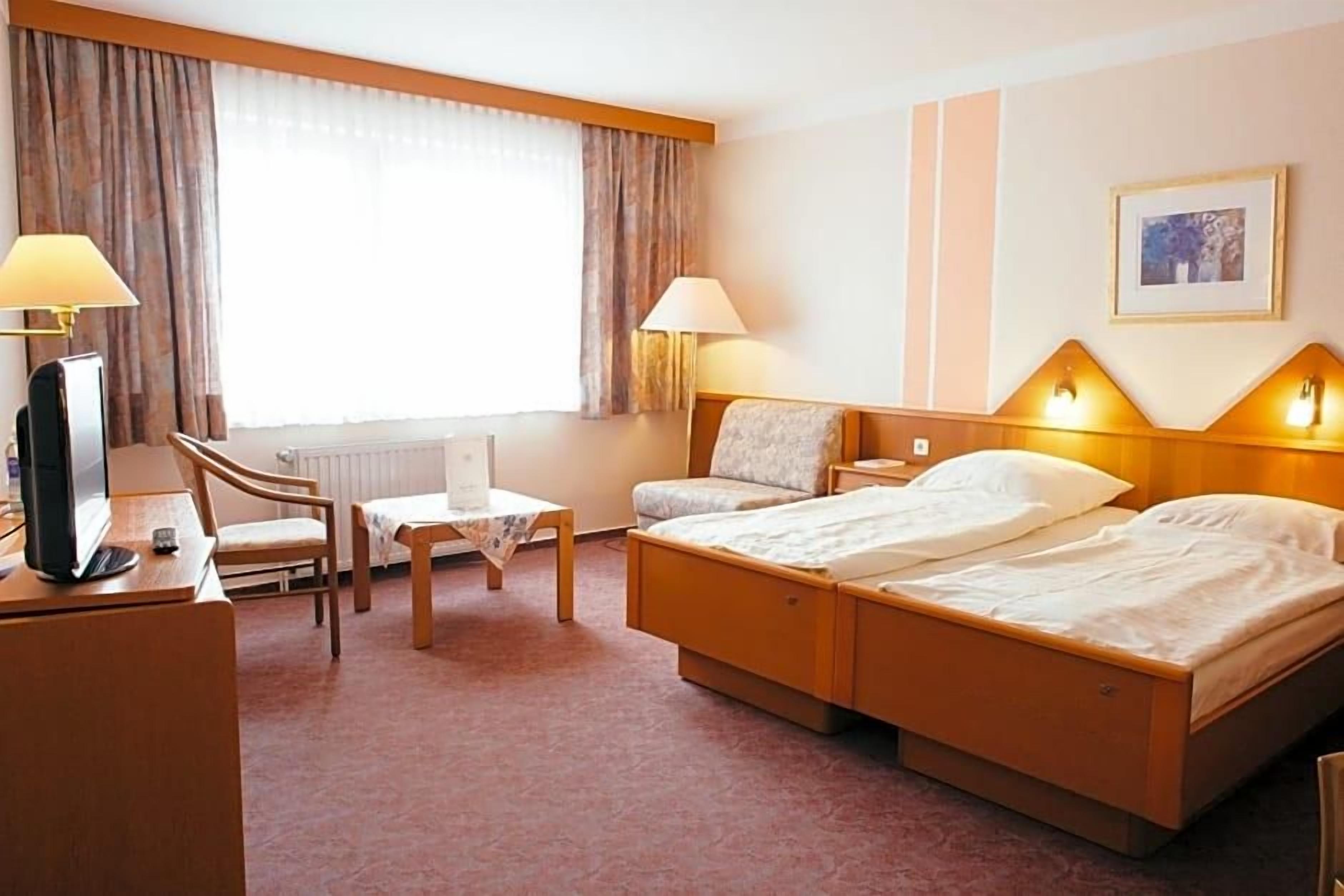 A hotel room with a double bed, a desk with a computer, a chair, a small table, and two bedside lamps.