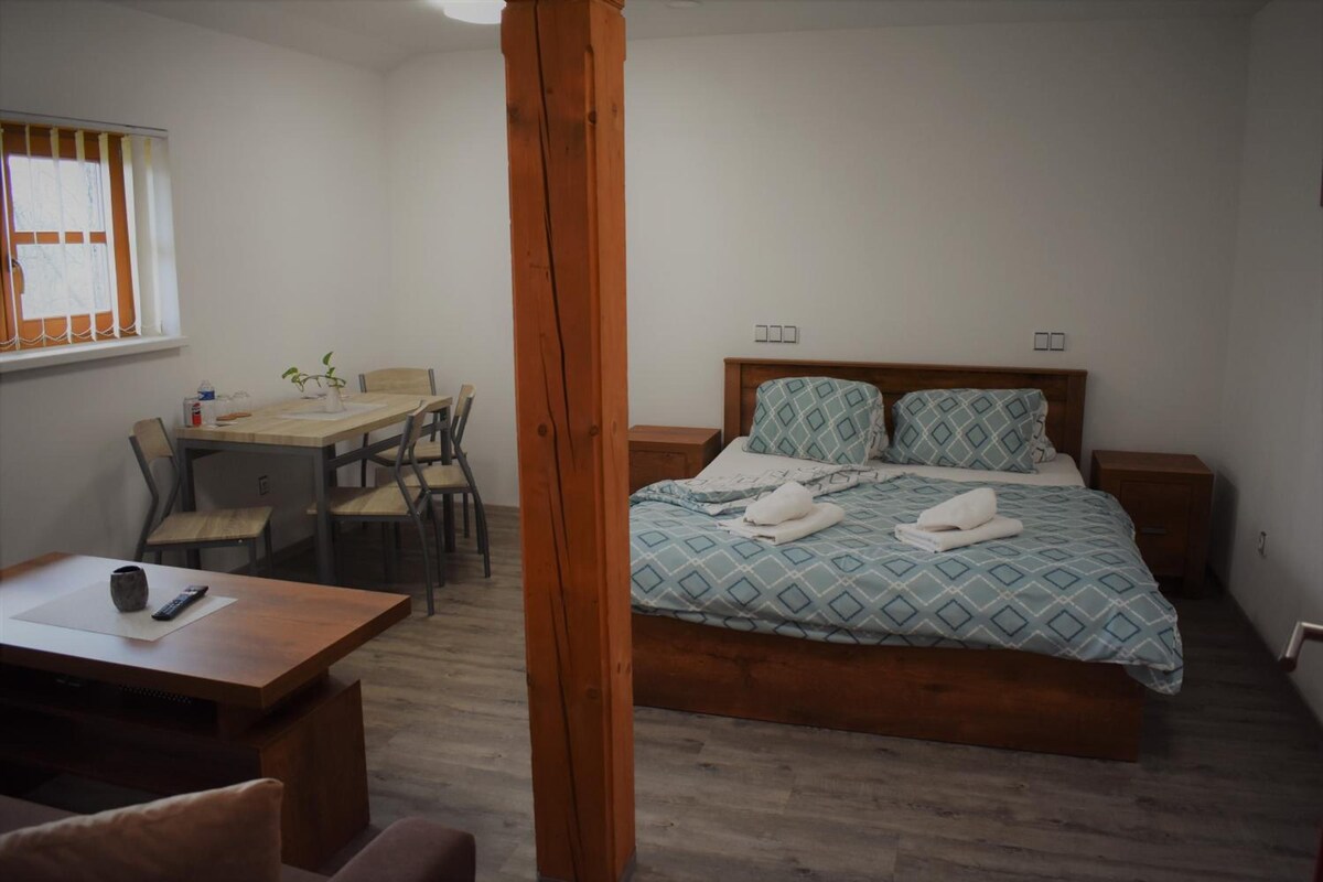 Un dormitorio con cama, mesitas de noche, una silla de madera, una pequeña mesa con un control remoto y una ventana con cortinas.