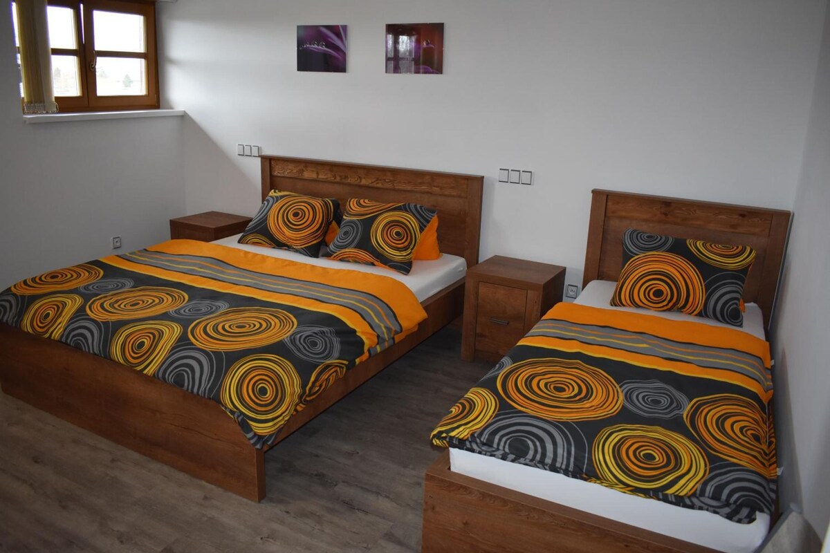 Una cama doble con colcha de estampado circular y almohadas a juego, cabecera de madera, mesita de noche y una ventana con persianas.