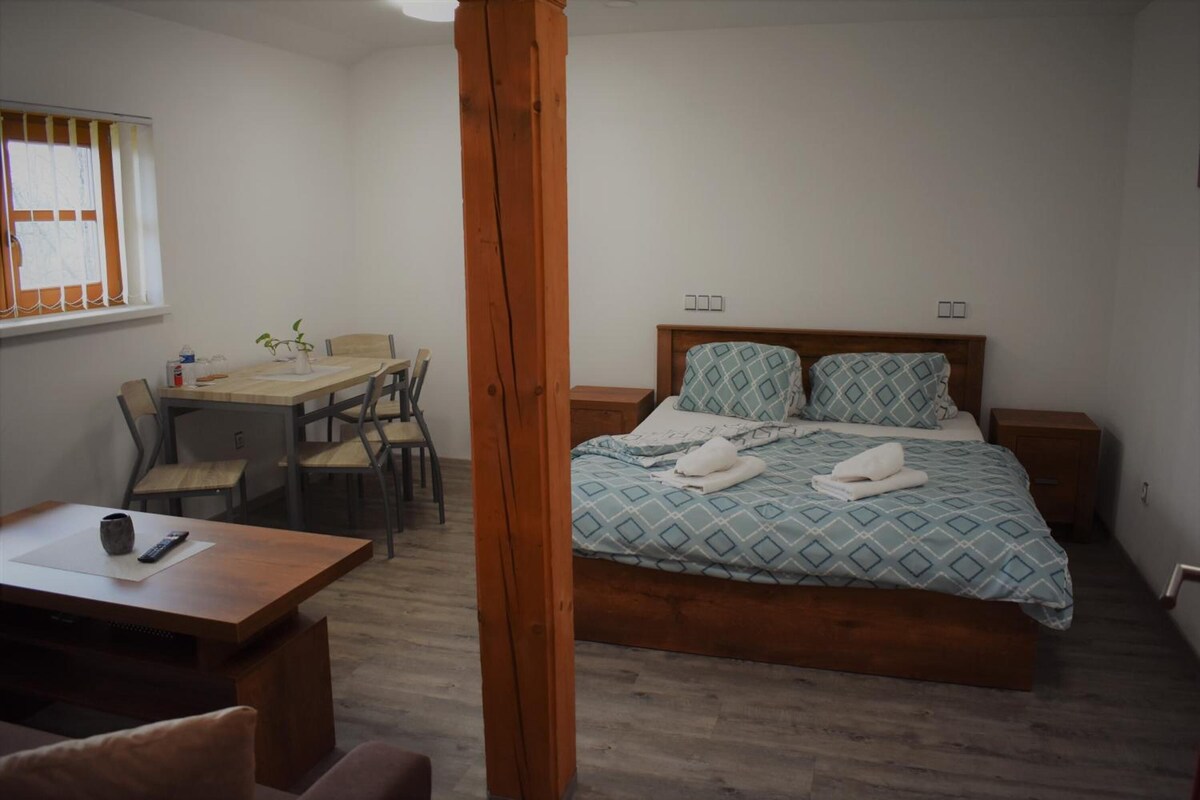 Un dormitorio con cama, mesitas de noche, una silla de madera, una pequeña mesa con un control remoto y una ventana con cortinas.