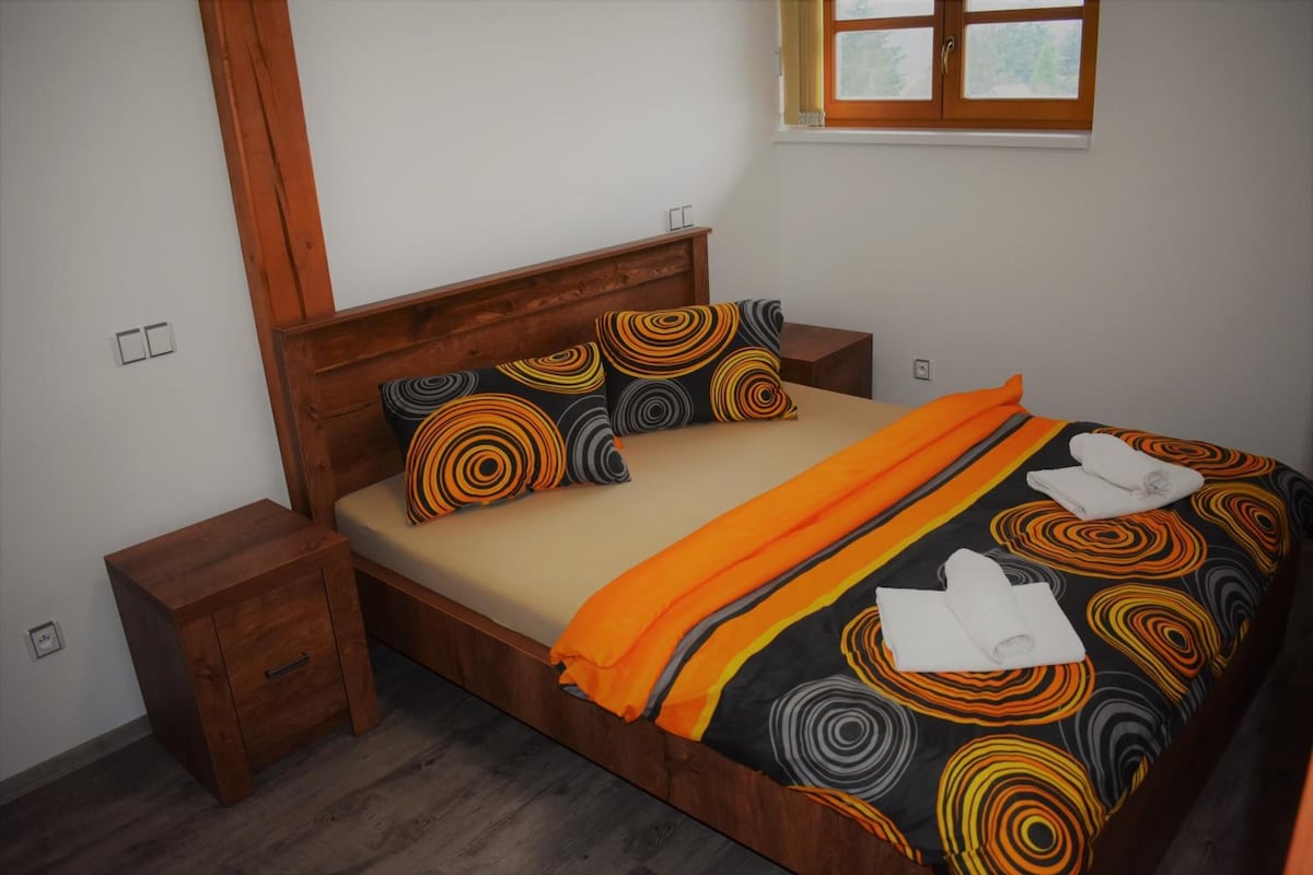 Un dormitorio con una cama de madera, dos mesitas de noche, un ventanal con cortinas y vistas a árboles.