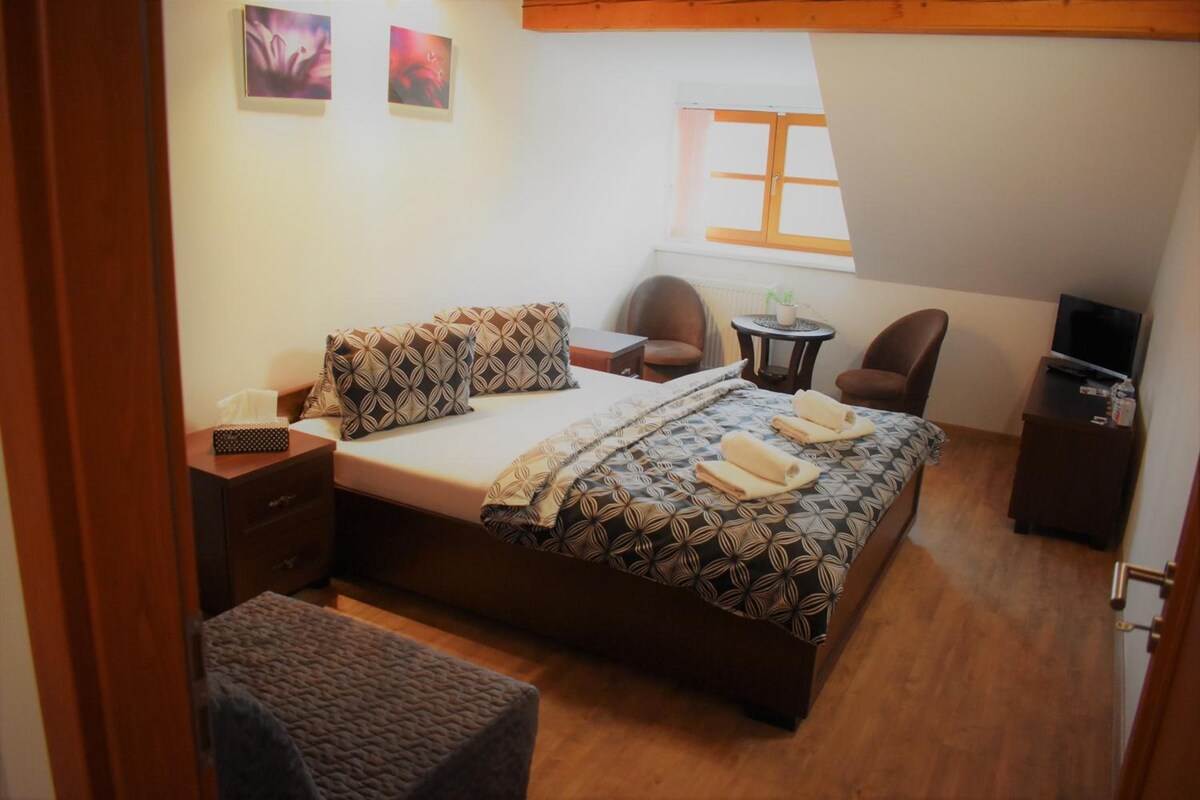 Un dormitorio con cama, dos sillas, una mesita, televisión y ventana.