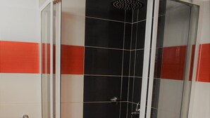 Ducha en esquina con mampara de vidrio, paredes con azulejos negros y rojos, y suelo blanco.