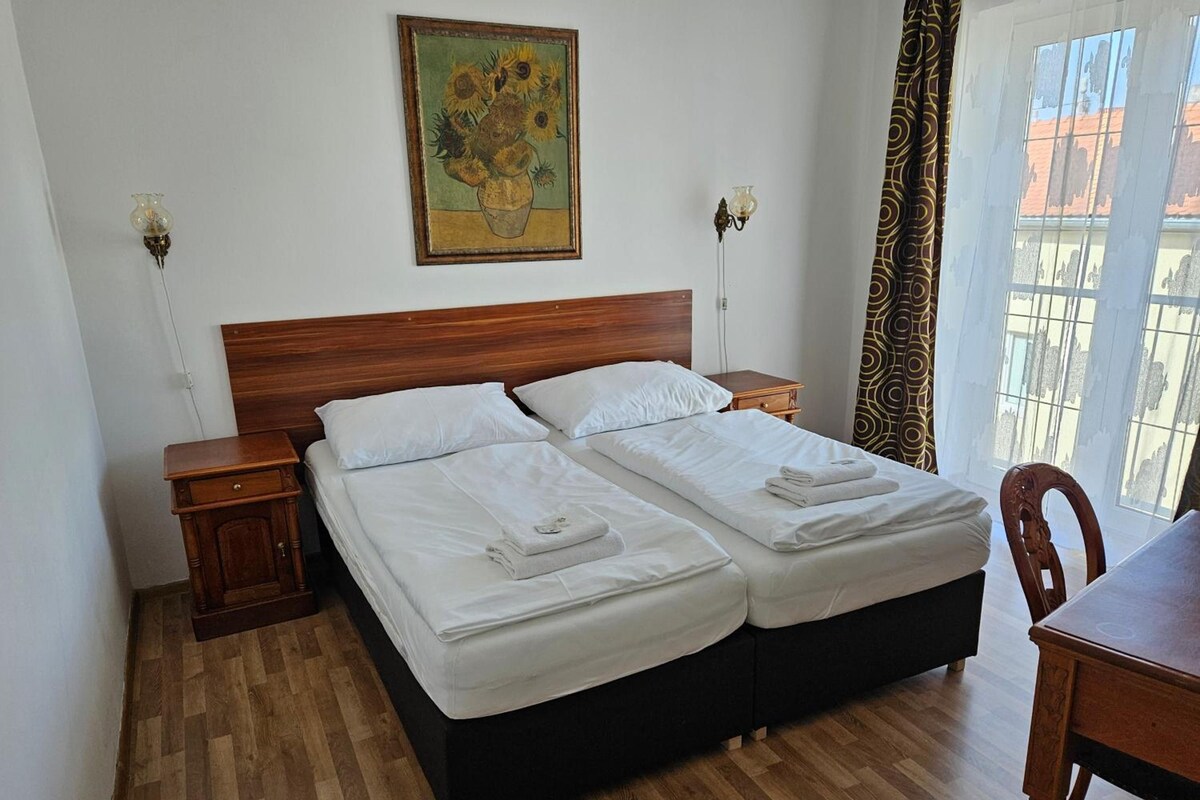 Una cama doble con ropa de cama blanca, un cabecero de madera, una mesita de noche pequeña, una lámpara de noche y un cuadro en la pared, además de una ventana con cortinas.