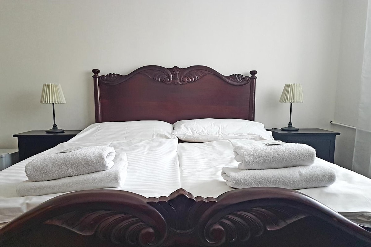 Una cama bien hecha con sábanas blancas y dos almohadas, flanqueada por dos mesitas de noche con lámparas, en una habitación con un cabecero de madera oscura.